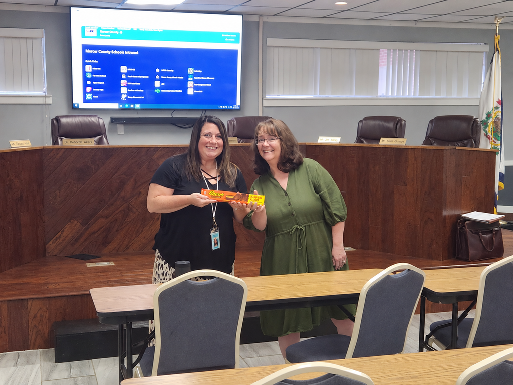 Two women stand in a meeting room, one holding a box, while a monitor displays text behind them.