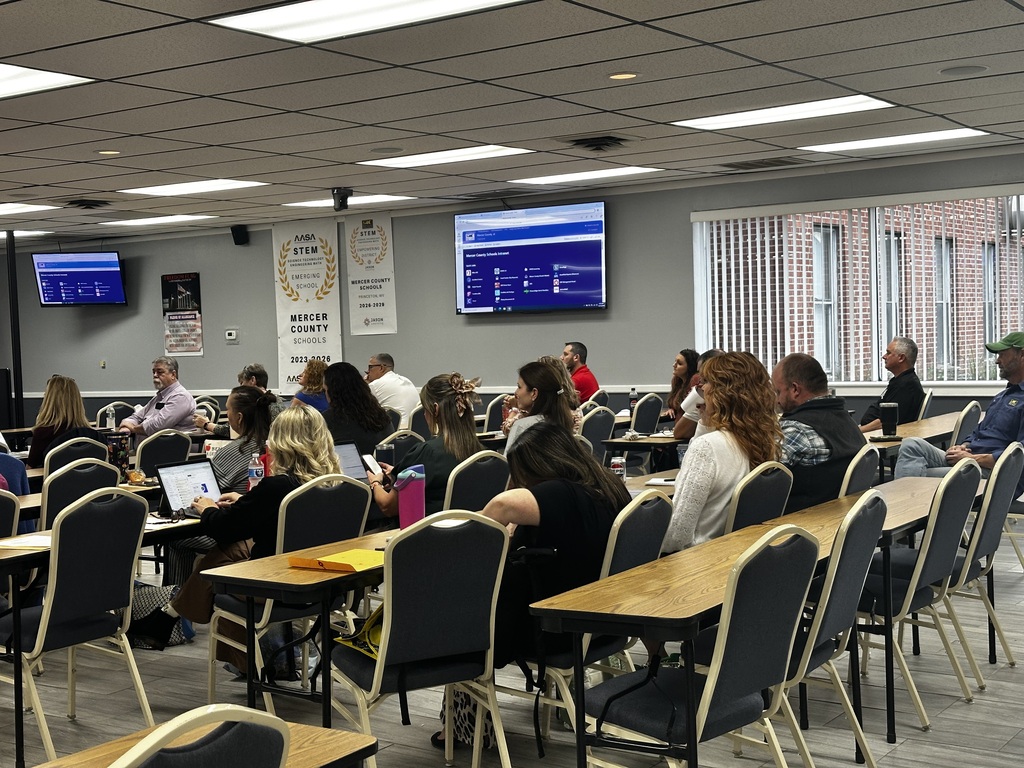 A large room with several rows of people in chairs. Some individuals use laptops. Monitors are on the wall.