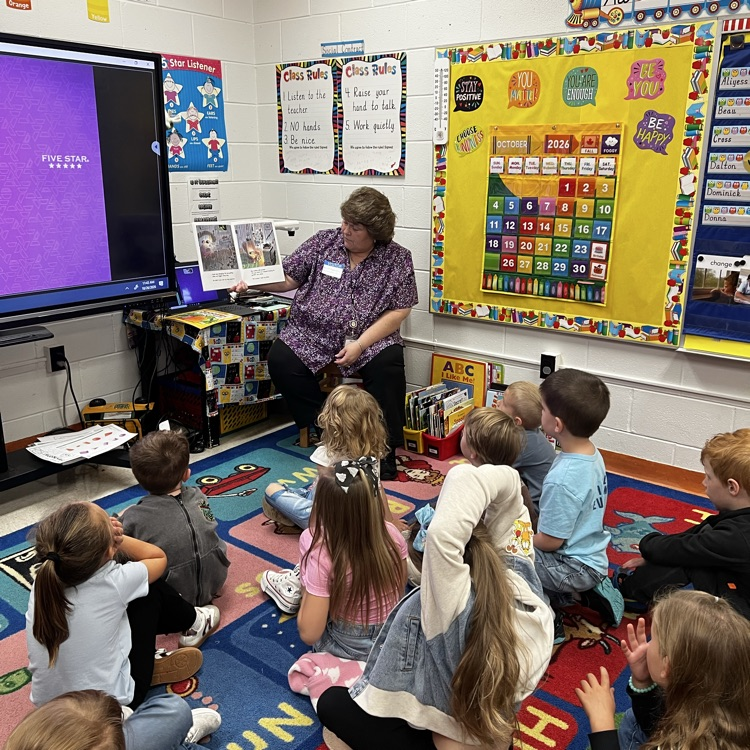 Woman in purple shirt reading a book to a group of children sitting on the floor.