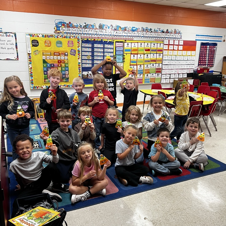 A classroom with children holding items. Some sit on the floor, others stand. Brightly colored charts on walls.