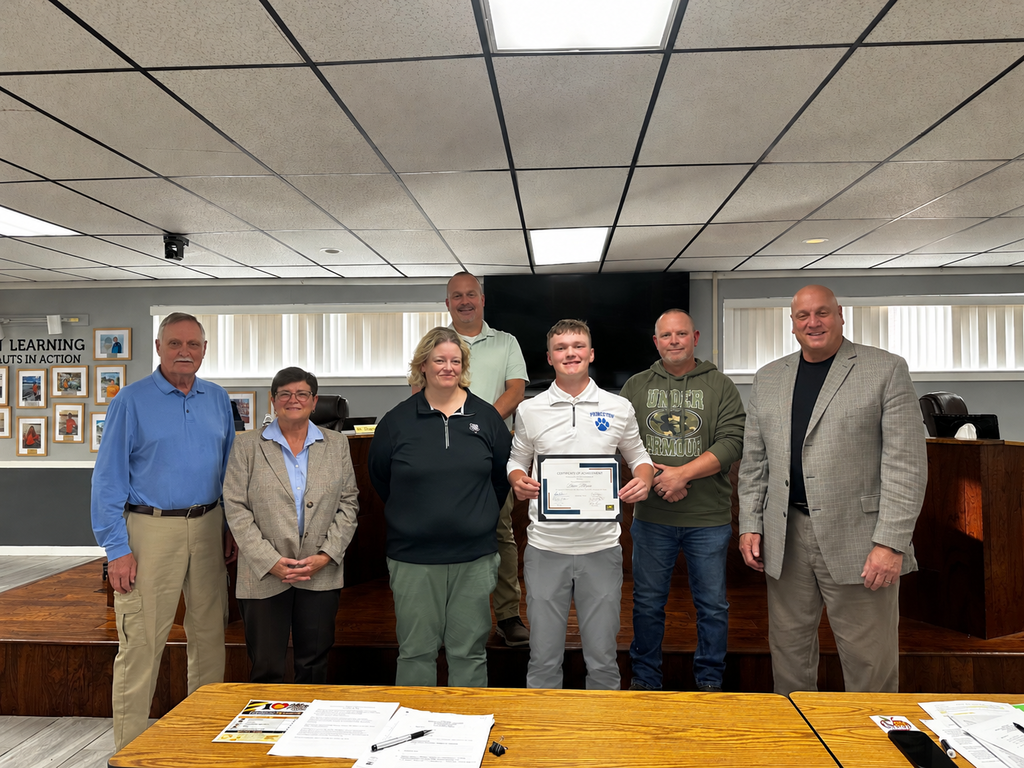 Six adults stand in an office with a wooden desk. One person holds a framed certificate.