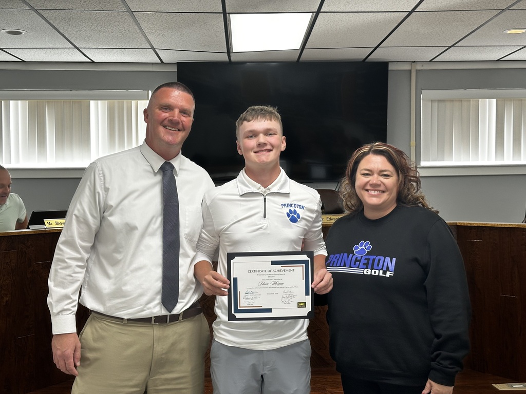 A man and a woman hold a certificate in front of a smiling boy. They stand in a room with a large monitor behind them.