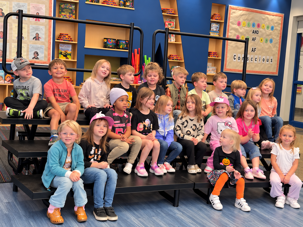 Group of children sitting on black platforms in a room with blue walls, shelves, and posters.