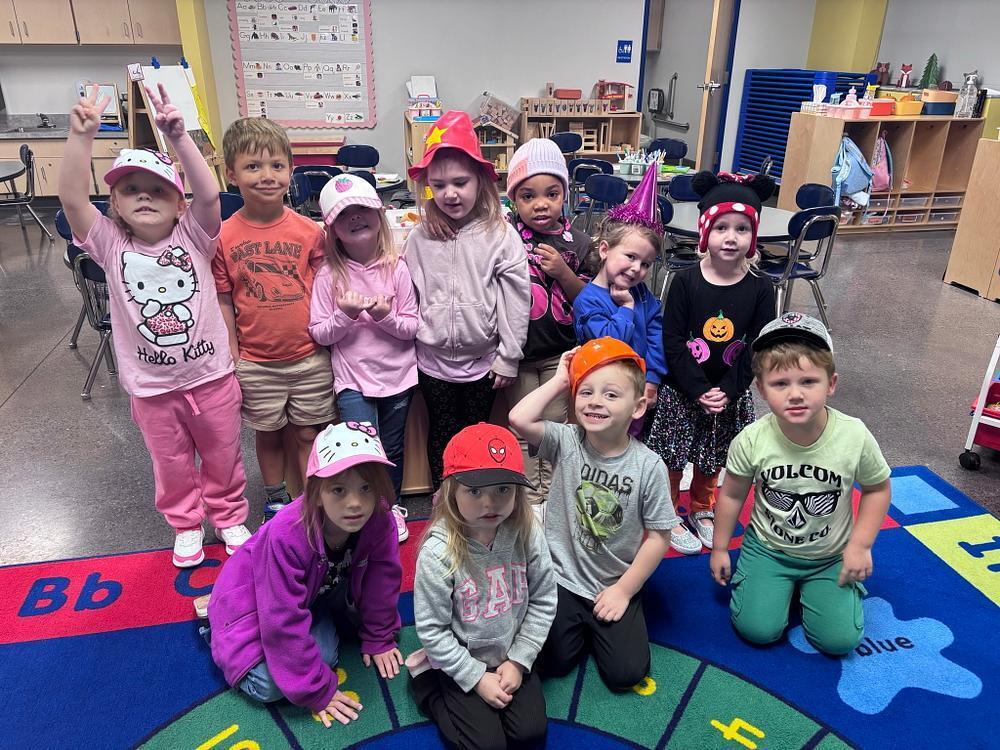 A classroom with a rug with alphabet letters. Nine children in hats crouch or stand on the rug and raise hands.