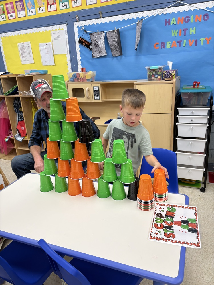 Two boys stack cups on a table. A blue wall with blue bulletin board is in the background.