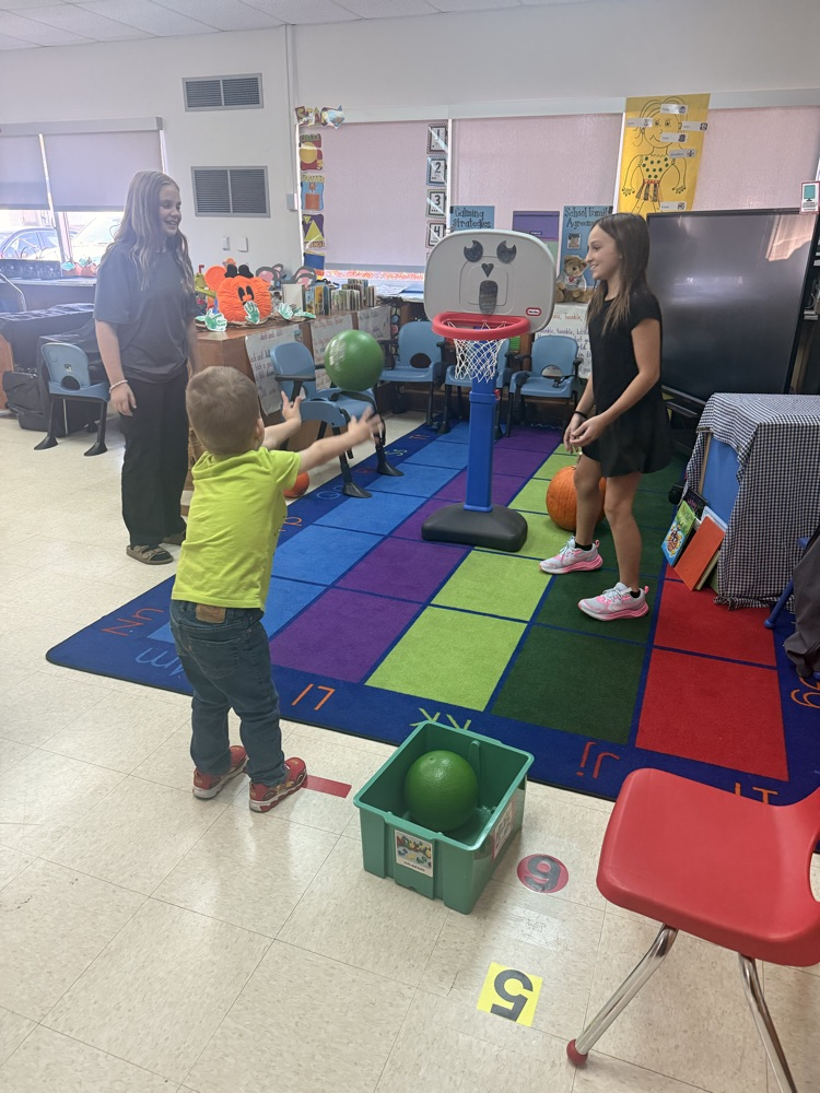 A classroom with a colorful rug; a boy throws a ball at a basketball hoop; two women watch.