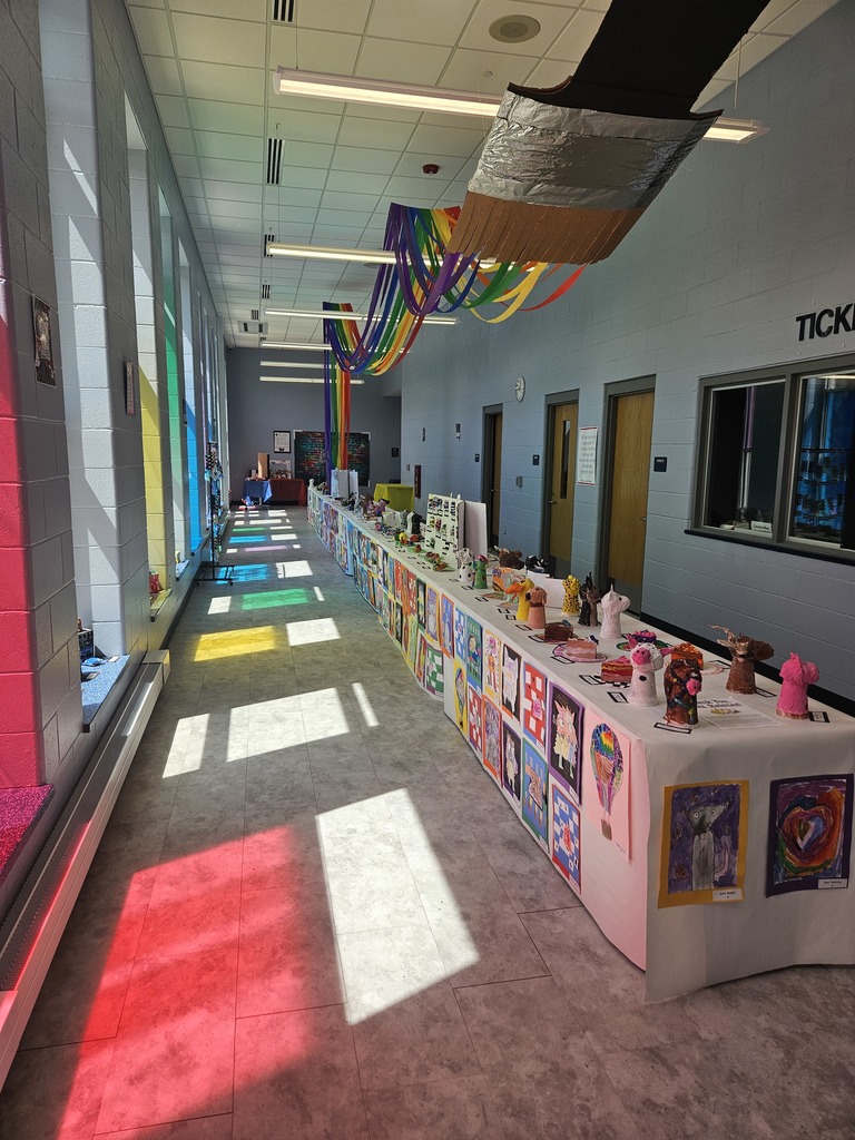 Elementary art projects showcased on a table with sunlight streaming through colored windows casting shadows on the floor. 