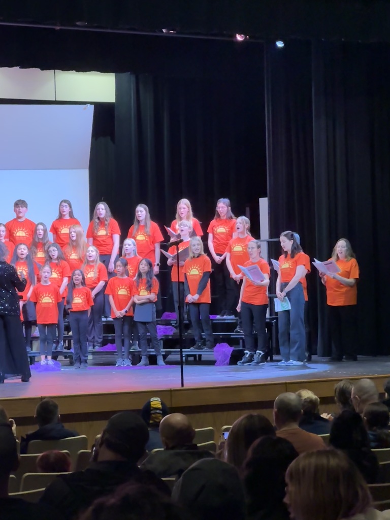 Students in orange shirts with a sun  on them singing on a stage