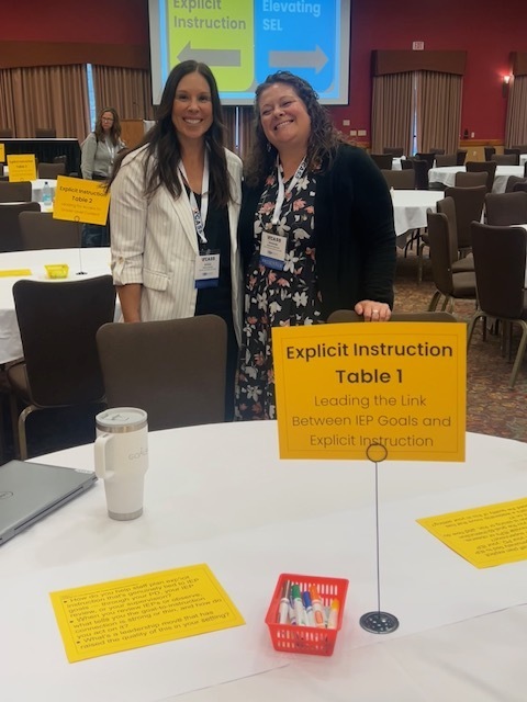 Two women standing by a table with a sign that says "Explicit Instruction Table 1"