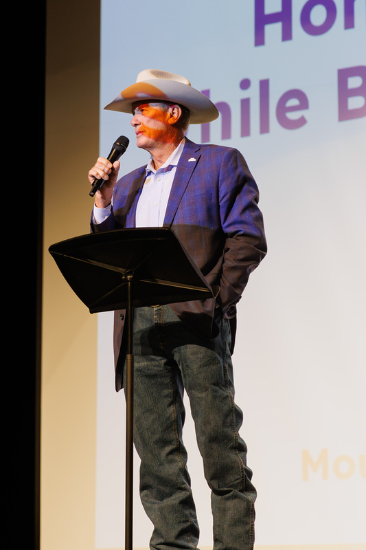 Mesa Mayor wearing a cowboy hat and blazer speaks into a microphone behind a music stand on stage.