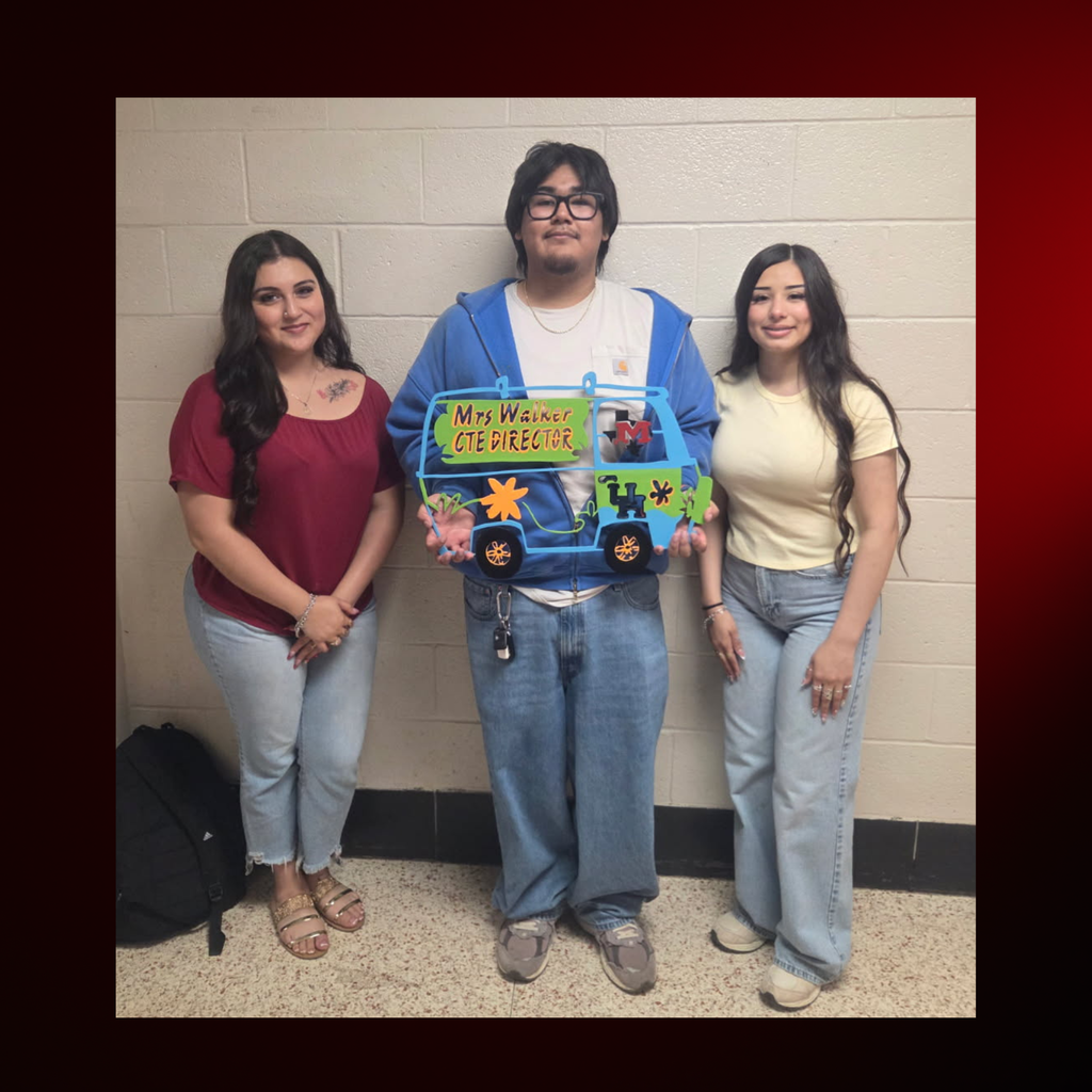 Three individuals pose for a photo, one holding a colorful artwork. They wear casual clothing and stand before a plain wall.