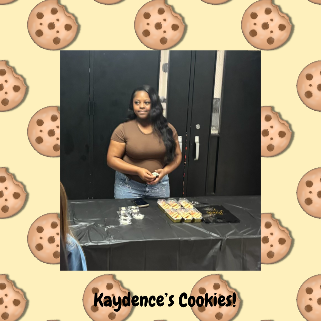 A woman stands behind a table with cookies, holding an item in her hands. The background features a patterned cookie design.