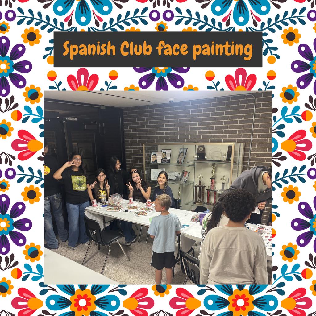 A group of people sit at a table, with some painting faces, in a room with a brick wall and decorations.