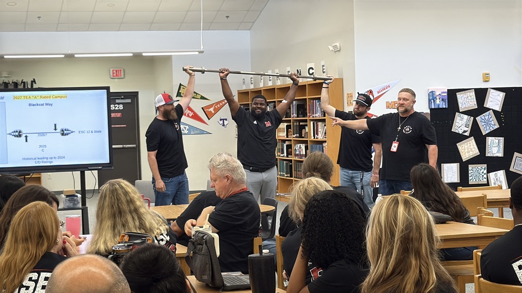 Several people in black T-shirts and baseball caps hold a metal bar above their heads in a classroom.