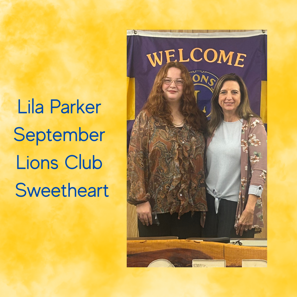 Two women stand behind a banner with "WELCOME" on it. One wears glasses and a patterned top, the other a floral jacket.