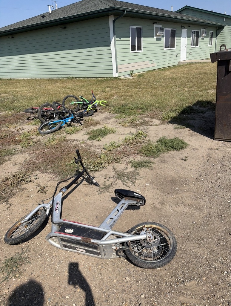 abandoned kids bikes laying in the dirt next to a dumpster in the summer sun