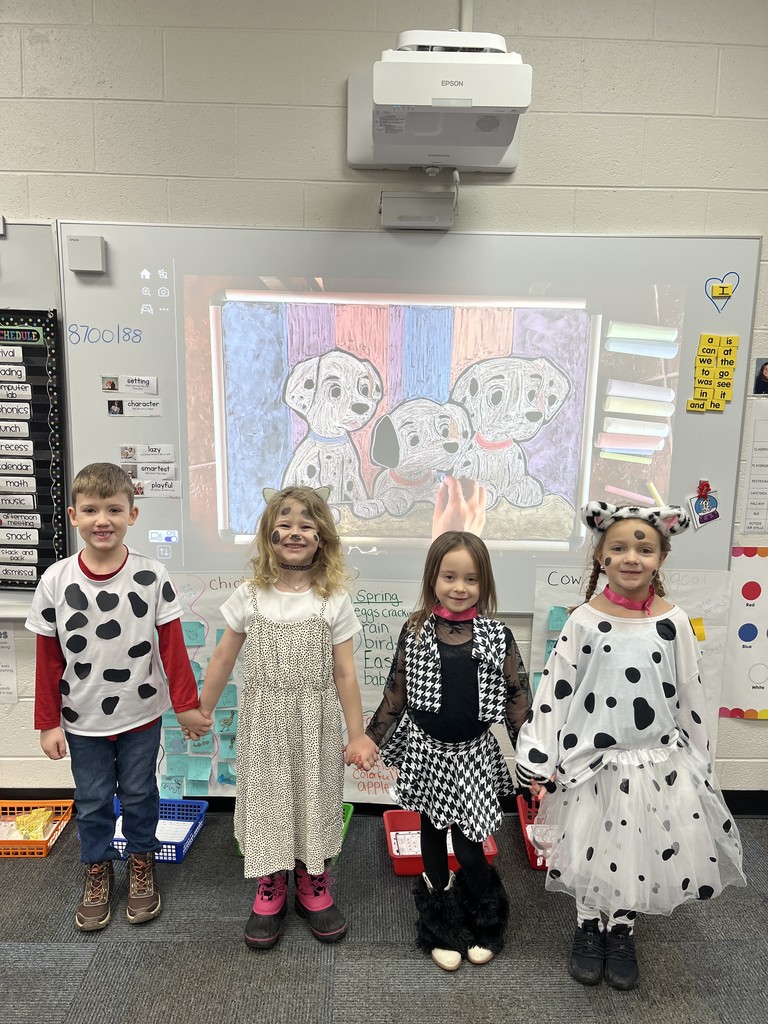 Four children dressed in costumes stand together in front of a classroom wall with a projector screen.