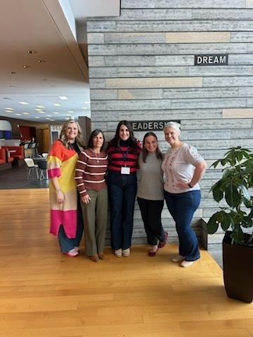 Five women stand in a hallway, smiling. Behind them is a wall with a stone design and signs reading "Dream" and "Leaders."