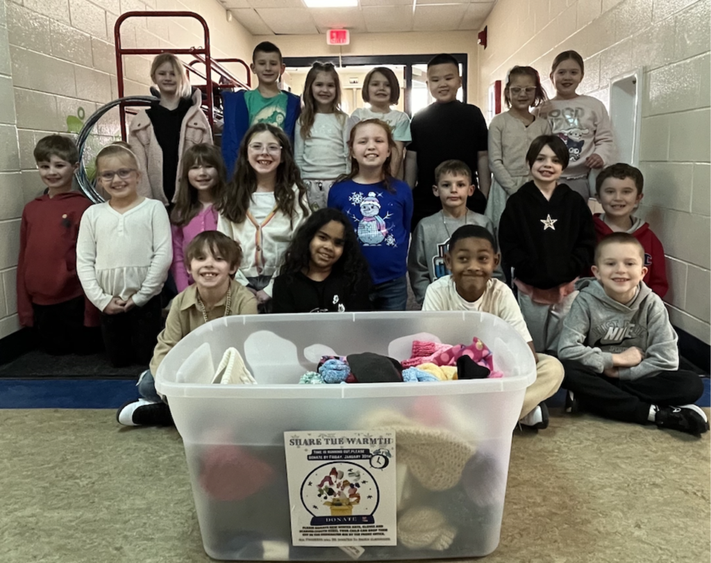 Kids pose for a group photo in a hallway. One sits in front of a box full of donated items.