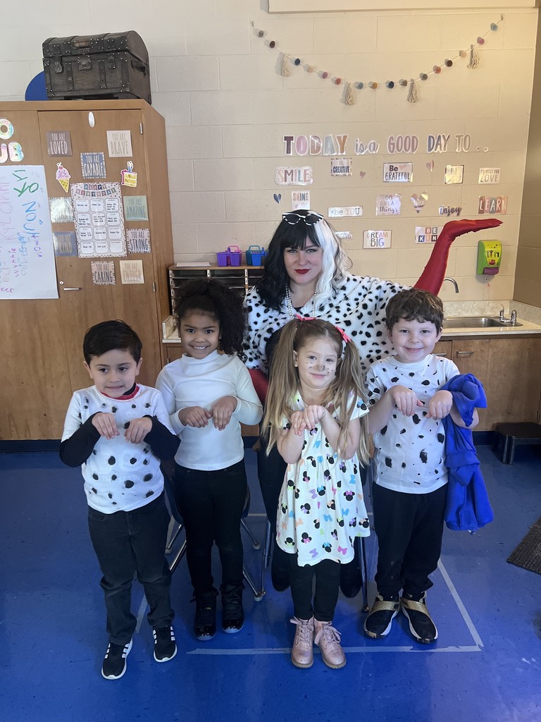 A group of children in polka-dot costumes poses with an adult in a similar costume. The room has a bulletin board, a wooden cabinet, and a sink.