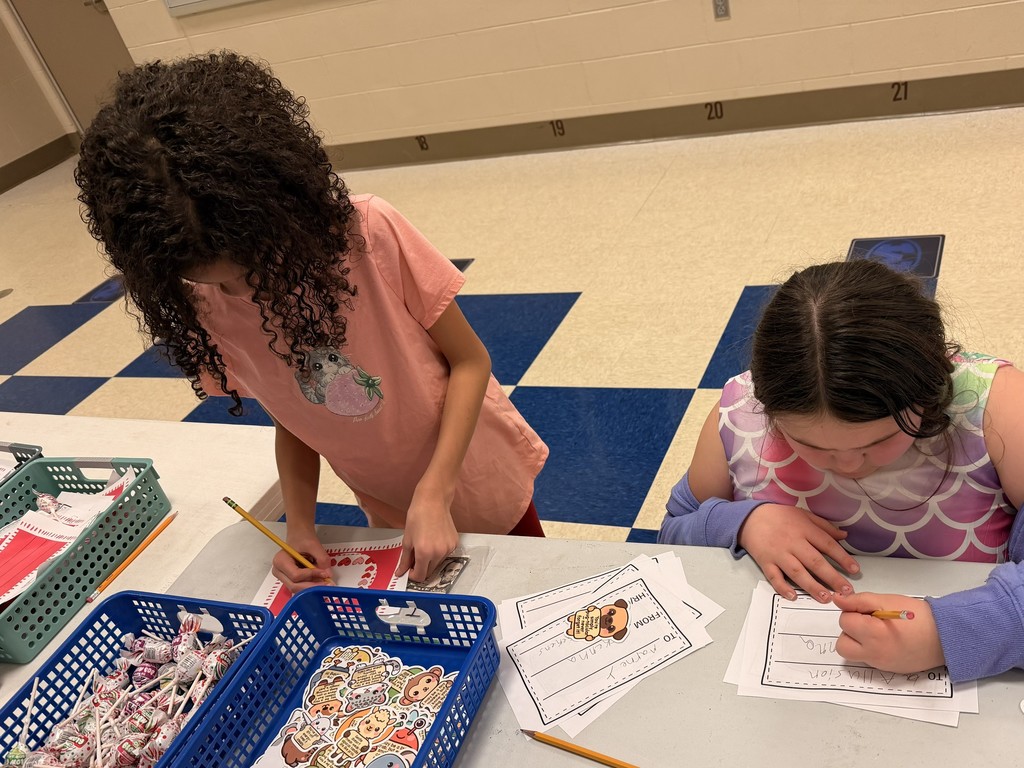 Two children in a classroom with a checkered floor. One with curly hair is bending over a desk with papers.