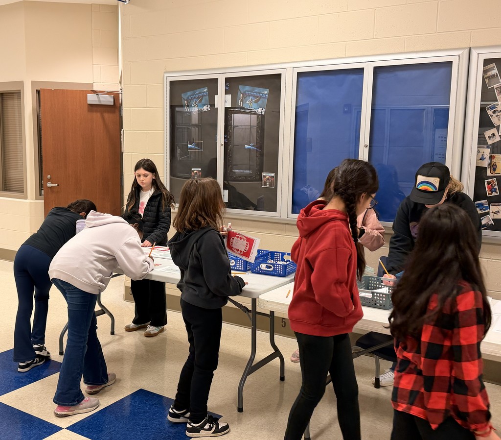 Students at tables, looking at objects. One girl bends over. Room has white walls and a blue checkered floor.