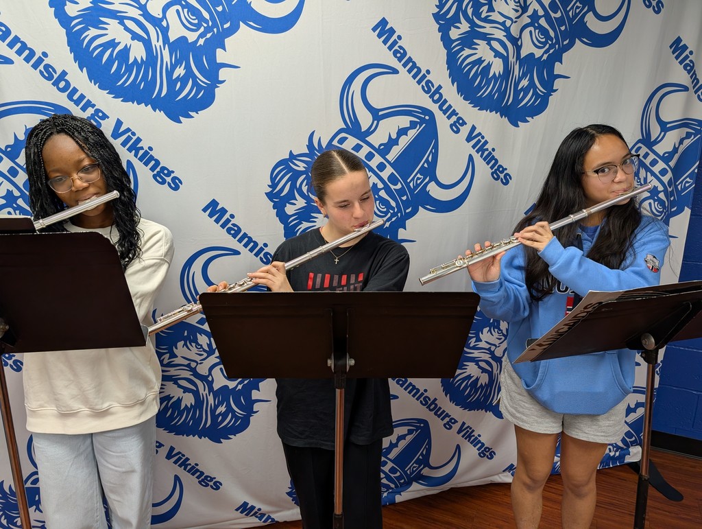 Three girls play flutes in front of a Miamisburg Vikings backdrop. One holds a music stand.
