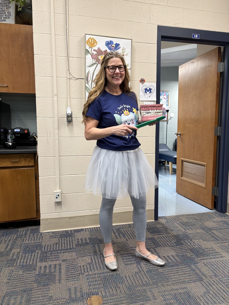 Woman in a blue shirt and tutu holding books stands in a room with wooden cabinets and open door.