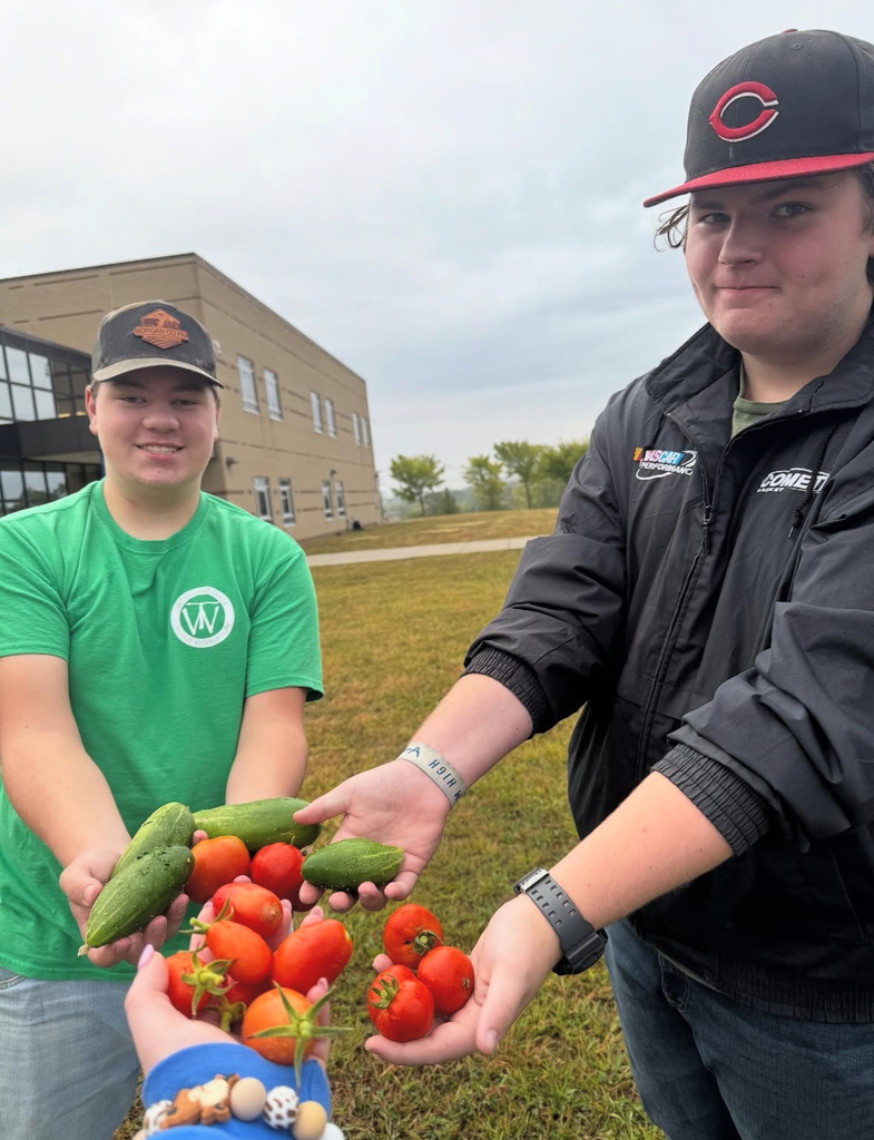 Two people holding vegetables. One wears a green shirt, and the other wears a black jacket. They are in front of a building.