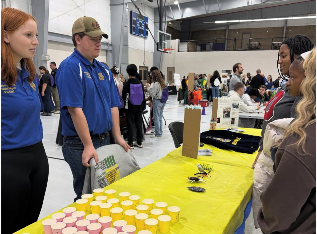 People gather at a table with yellow tablecloths, cups, and various items, while a man in a blue shirt talks.