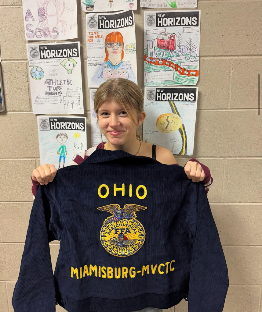 Person in a classroom, holding up a navy blue jacket with "OHIO" and "MIAMISBURG-MVCTC" in yellow letters.