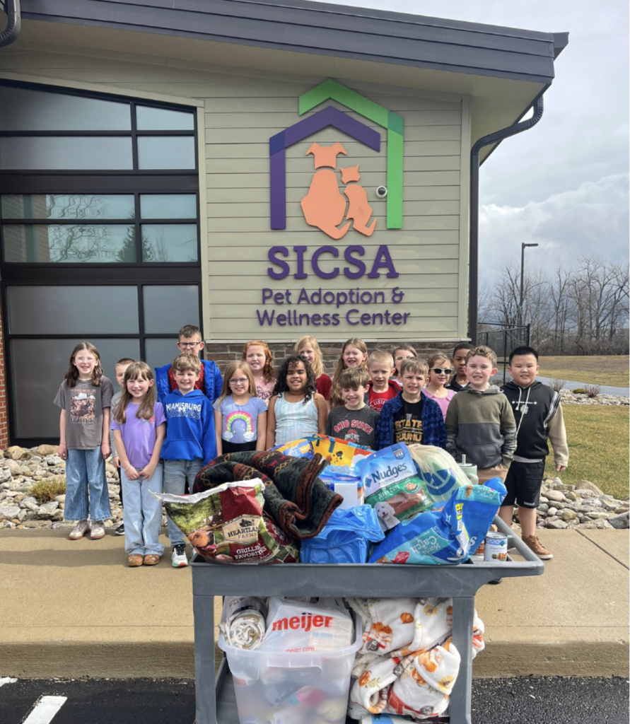 Children stand in front of a building with a sign for SICCSA. They pose with a cart full of goods.
