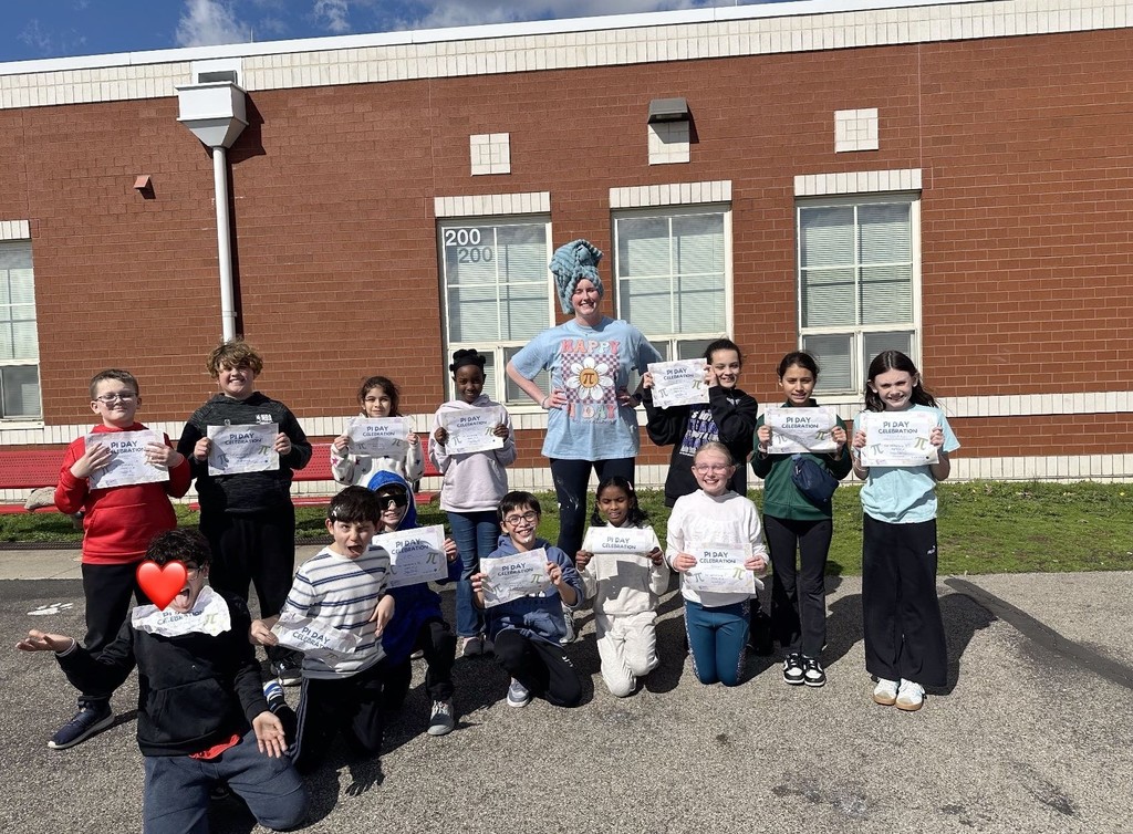 A group of children, some wearing costumes, stand in a line and hold certificates in front of a brick building.