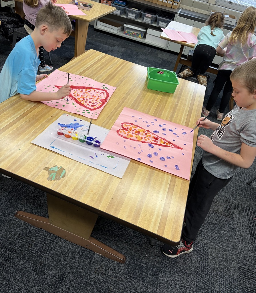 Children seated at a table in a classroom. One child dips paintbrushes into colored paint.