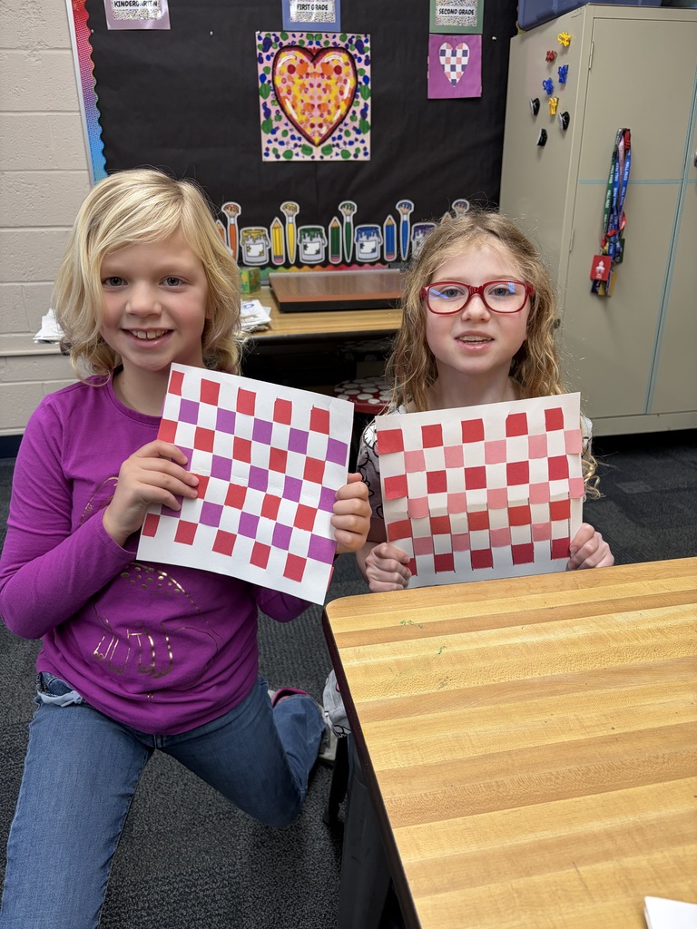 Two girls hold up checkered paper in a classroom with a bulletin board in the background.