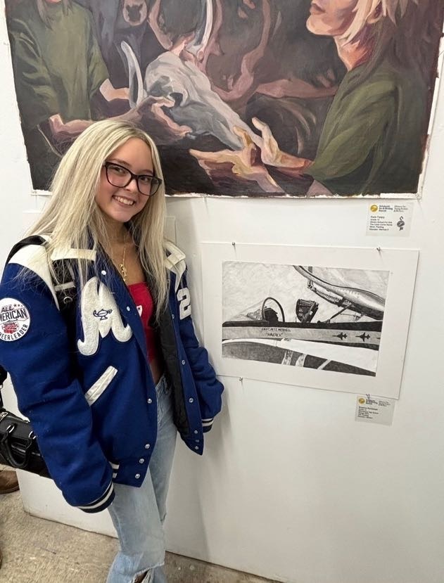 Woman with glasses and a blue jacket stands next to an art exhibit on a white wall.