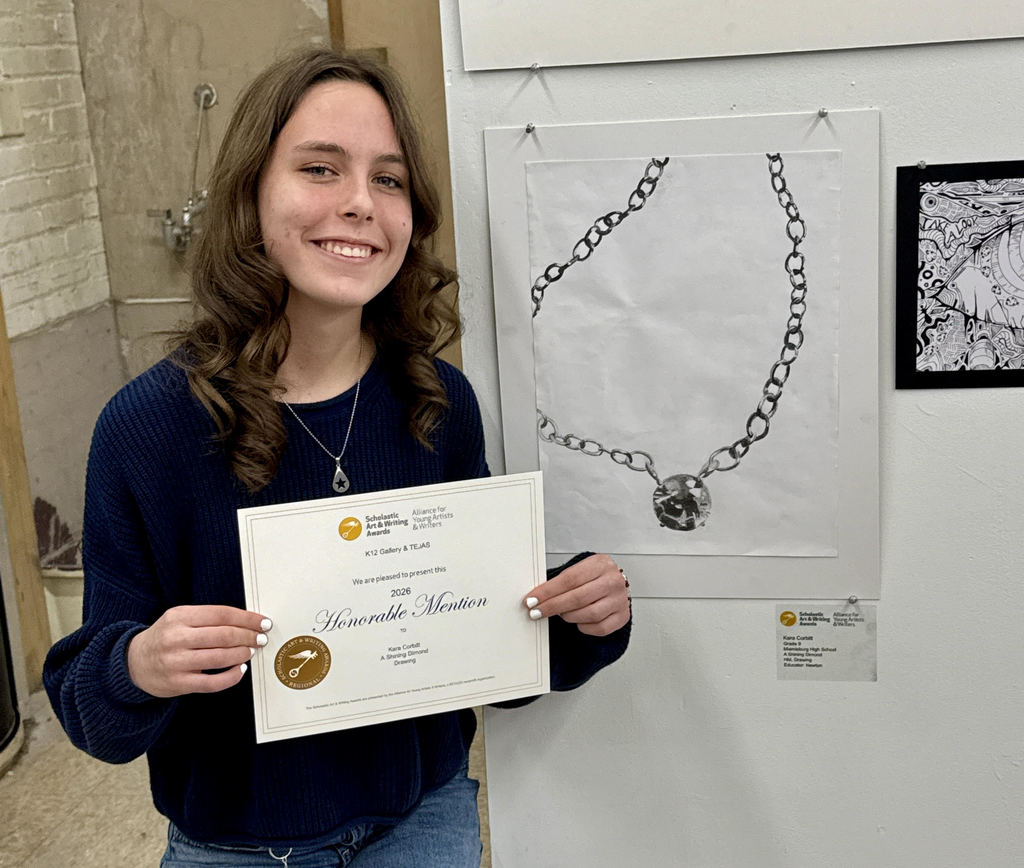 Person holding an award certificate in front of a white wall. Behind, two framed artwork displays.