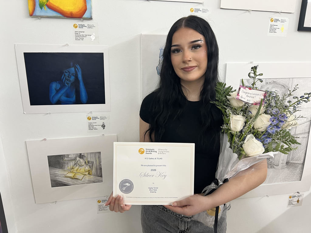 Person holds certificate and flowers in front of wall with framed pictures. Background includes digital displays.