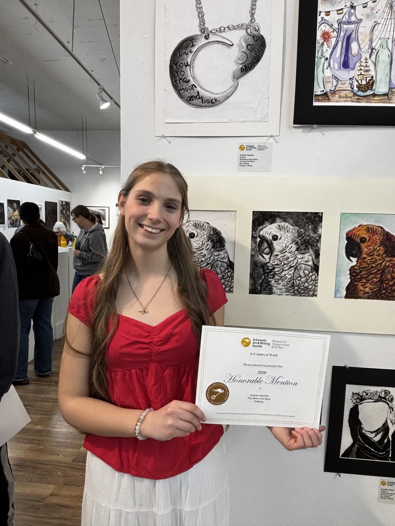 Woman in red shirt holding framed certificate in front of art gallery with framed artwork on wall.