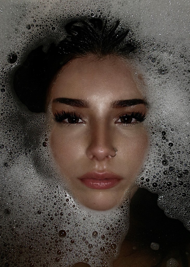 A woman with dark hair is partially submerged in bubbles, gazing directly at the camera. She has makeup on.