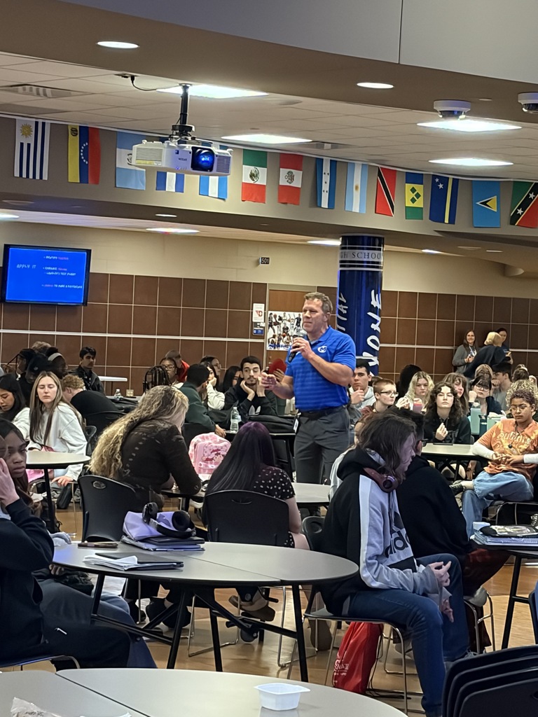 A man in a blue shirt stands at a podium in a classroom with many students. Flags hang from the ceiling.