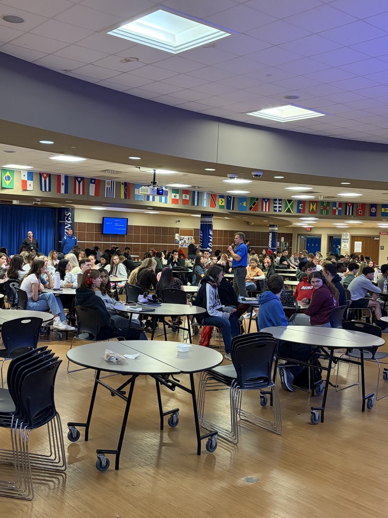 Large room with tables and chairs. Many people seated at tables, some standing. Flags hang on the wall, and a screen is visible.
