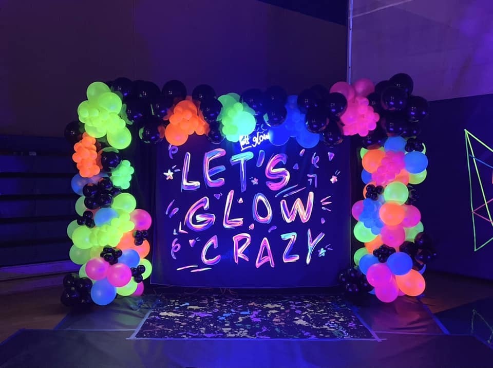 Neon lights illuminate a backdrop of balloons in various colors. A black frame surrounds the balloons, and a sign reads "Let's Glow Crazy."