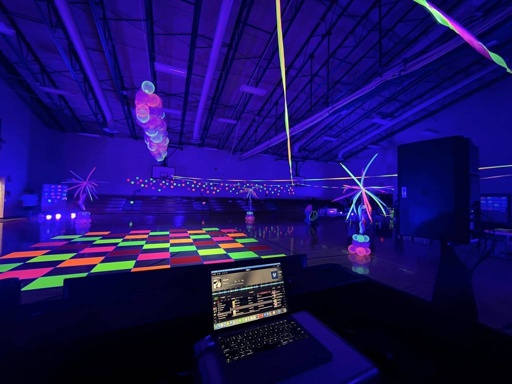 A dark room with multicolored neon lights and a checkered dance floor. A laptop displays on a table.