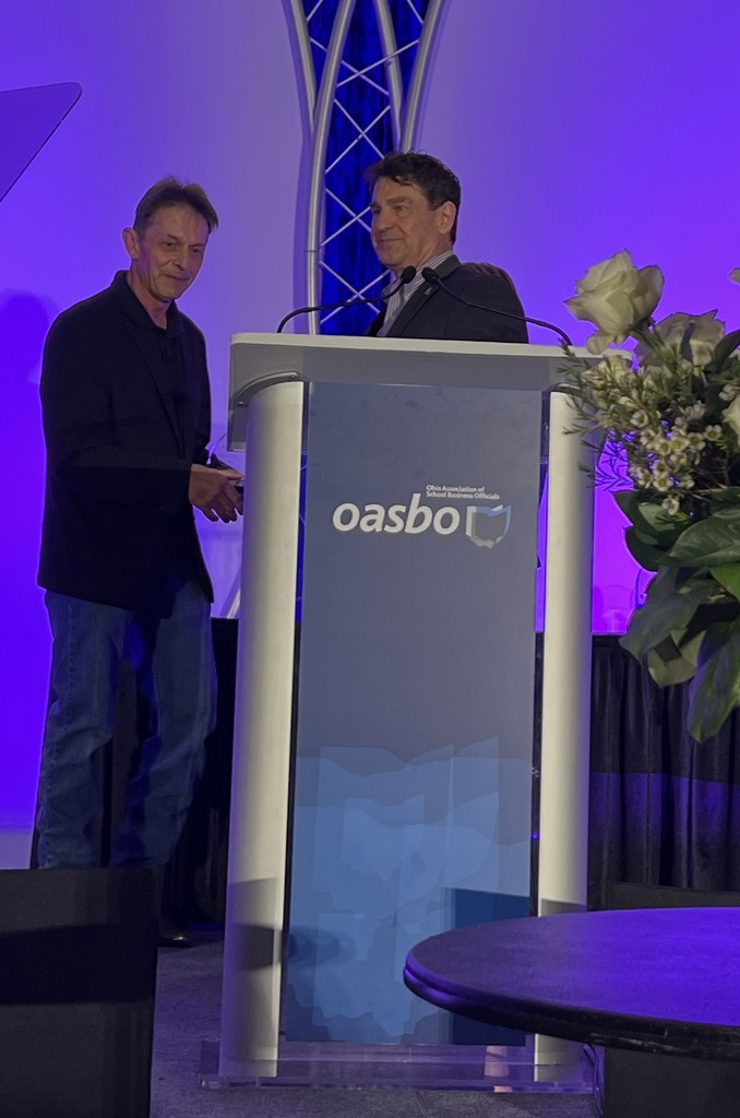 Two men standing at a podium with microphones, one smiling. A white rose arrangement is on the podium.