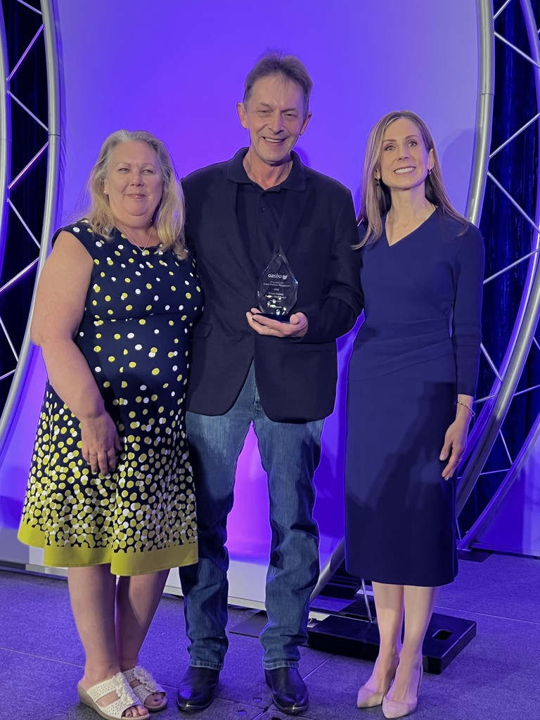 Three people stand on a stage with one holding an award. Purple lighting and a metal structure in the background.