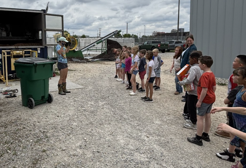 A group of children stands in a row in front of a recycling facility, with one woman speaking to them.