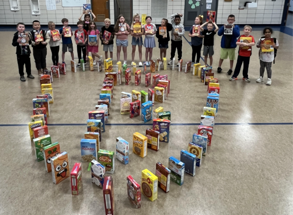 A group of children holding boxes of cereal arranged in a heart shape on a gym floor.