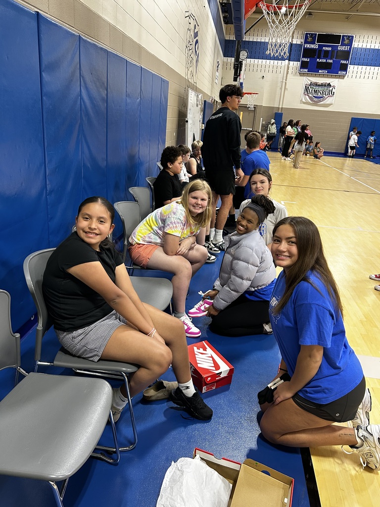 Group of young people sitting and kneeling on a basketball court. Some are wearing athletic gear.