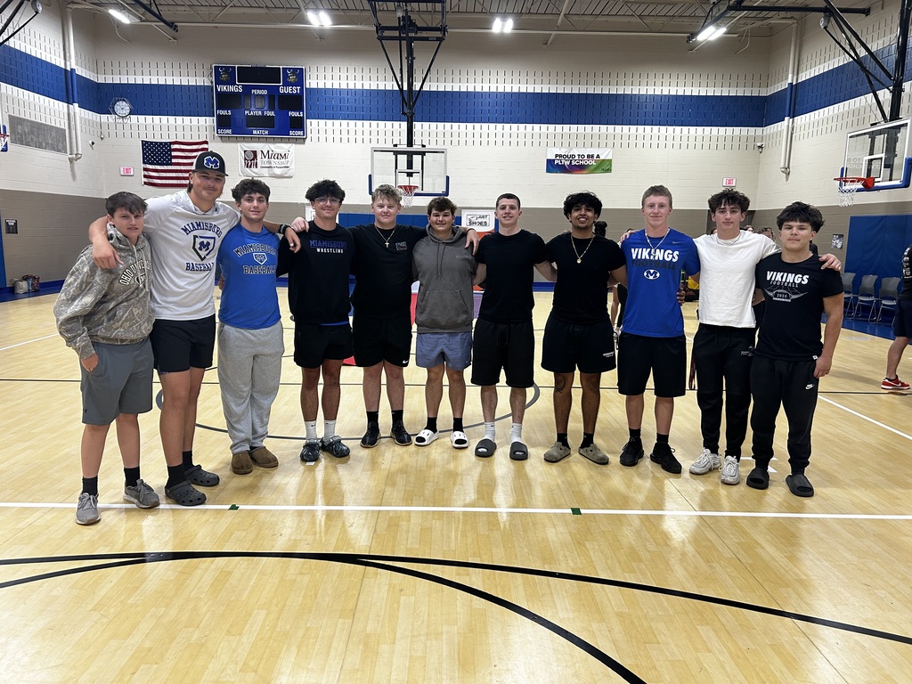 A group of young men in sports attire stands on a basketball court. Some wear hats.